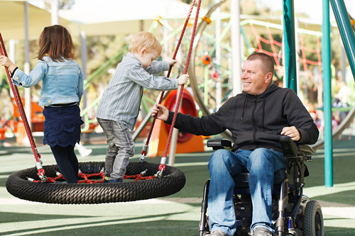 A man in a wheelchair assists children playing on a tire swing at a playground.